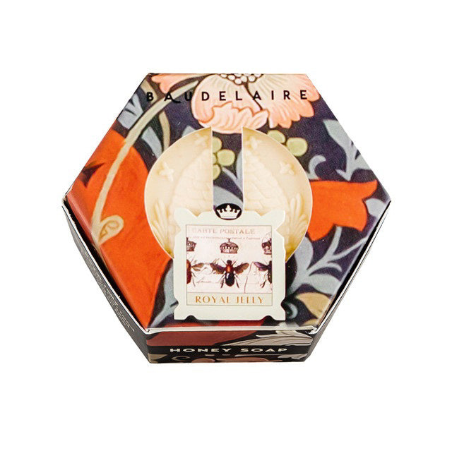 The BAUDELAIRE - ROYAL JELLY HONEY SOAP HEX BOX 3.5 OZ features a hexagonal box adorned with a floral design and &quot;Baudelaire&quot; elegantly written on top. At the center, a circular label showcases a vintage bee illustration with the text &quot;Royal Jelly Soap.&quot; Inside, you’ll find honey-colored soap with an embossed design that promises to hydrate your skin for a glowing complexion.