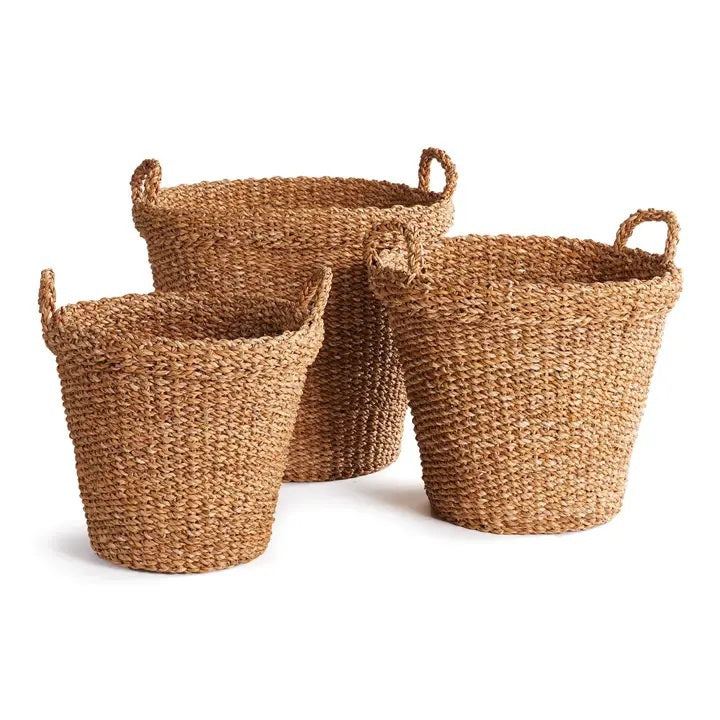 A NAPA HOME AND GARDEN Seagrass Tapered Basket with Handles and Cuff (Medium) sits on a woven rug, holding tall decorative green grass, with a chair and colorful painting in the background.