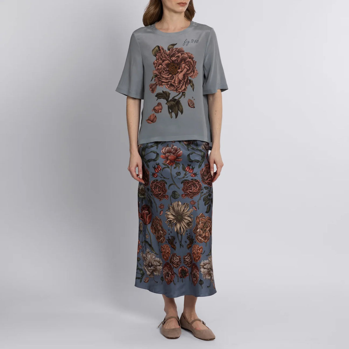 A person is seen from the back wearing a SABINA SAVAGE RING A RING O&#39; ROSES SILK BIAS SKIRT, featuring English garden flowers in red, brown, and cream on gray-blue. They pair it elegantly with tan flat slip-on shoes.