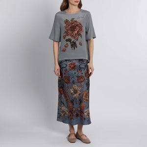 A person is seen from the back wearing a SABINA SAVAGE RING A RING O' ROSES SILK BIAS SKIRT, featuring English garden flowers in red, brown, and cream on gray-blue. They pair it elegantly with tan flat slip-on shoes.