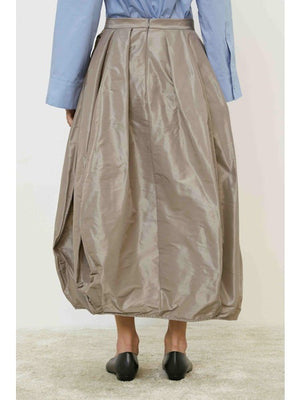 A person wears the MEIMEIJ Taffeta Balloon Skirt in shiny taupe, paired with a light blue long-sleeve shirt and black flats, standing on a light carpet against a white background.