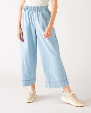 Wearing MERSEA - SAMMIE WIDE LEG PANT in light blue chambray with a pull-on waistband and front patch pockets, styled with a light top and cream sneakers, the person stands against a plain background.