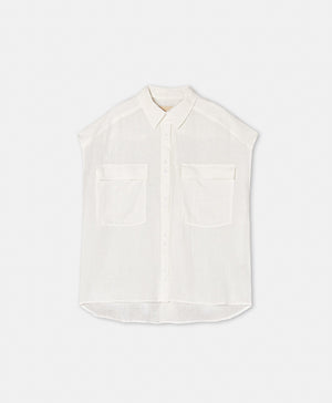 A woman with long blonde hair models the MOMONI SLEEVELESS LINEN SHIRT, a loose-fitting, garment-dyed white button-up with two front pockets, paired with black pants, standing against a plain light background.