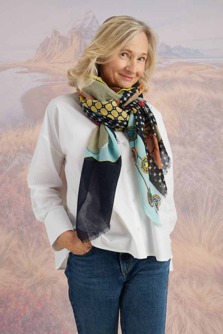 A woman with long blonde hair, in a light blue shirt and dark pants, smiles in front of a scenic backdrop, draping the elegant, hand-embroidered STORIATIPIC - ASTER WOOL SCARF around her shoulders.