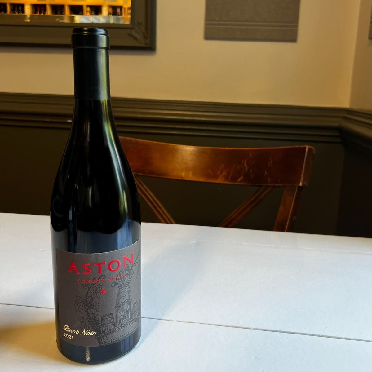 A bottle of ASTON ESTATE PINOT NOIR 2021 with a dark label featuring red text and a wine-themed illustration, sitting on a white table.