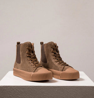 A pair of DANIELLA LEHAVI - OSAKA HIGH TOP SNEAKERS with a contemporary design, featuring brown suede uppers and rugged rubber soles, are placed side by side on a white pedestal against a plain beige background. These comfortable high-top sneakers boast a lace-up design and ribbed toe caps for added durability.