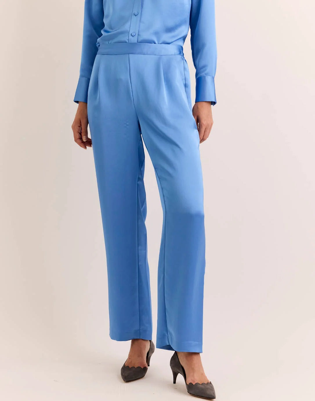 Model wears the HINSON WU - HOSTESS LIQUID SATIN PANT IN SKY BLUE by HINSON WU, featuring a relaxed straight leg and elastic waistband in luminous, light-catching sky blue satin. Styled with the matching long-sleeve top, they are pictured from the back against a neutral backdrop and complete the look with gray pointed-toe shoes.