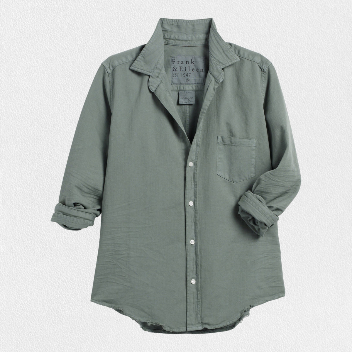 The FRANK AND EILEEN - BARRY TAILORED BUTTON UP SHIRT IN FAMOUS DENIM THYME, displayed against a white background, is a light gray-green classic shirt. Crafted from Liberty Fabric, it features a front pocket on the left side and long sleeves rolled up to the elbows. The shirt has a slightly curved hemline, a pointed collar, and a tailored fit. The inside label reads &quot;Frank &amp; Eileen EST. 1947.
