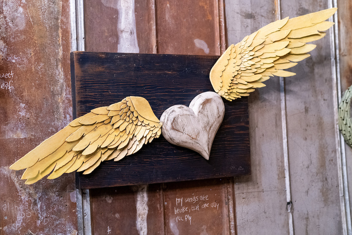 Wooden heart with large yellow wings attached, mounted on a dark rectangular board against a rustic metal wall. Handwritten text is faintly visible on the wall nearby.