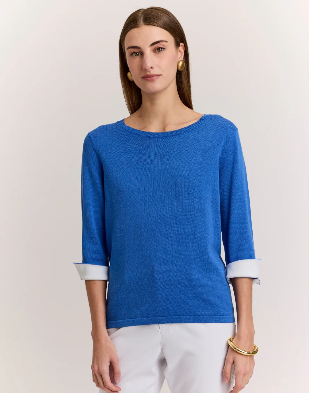 A woman with straight brown hair and gold earrings wears the HINSON WU TYLER 3/4 SLEEVE SWEATER in bright blue, styled with rolled cuffs, white pants, and a gold bracelet, standing against a neutral background.