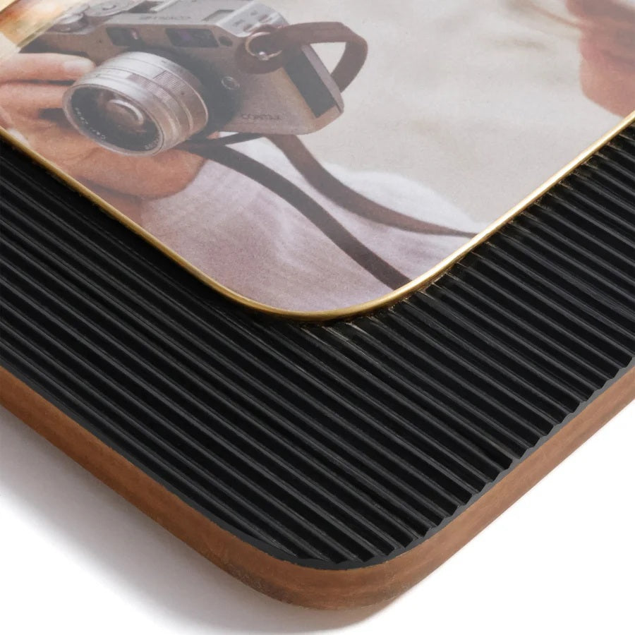 Two ZODAX BLACK RESIN RIBBED PHOTO FRAMES WITH BRASS ACCENT (5x7) display photos of a person in white, one with a camera. Each frame rests on beige boxes against a light blue background.