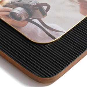 Two ZODAX BLACK RESIN RIBBED PHOTO FRAMES WITH BRASS ACCENT (5x7) display photos of a person in white, one with a camera. Each frame rests on beige boxes against a light blue background.