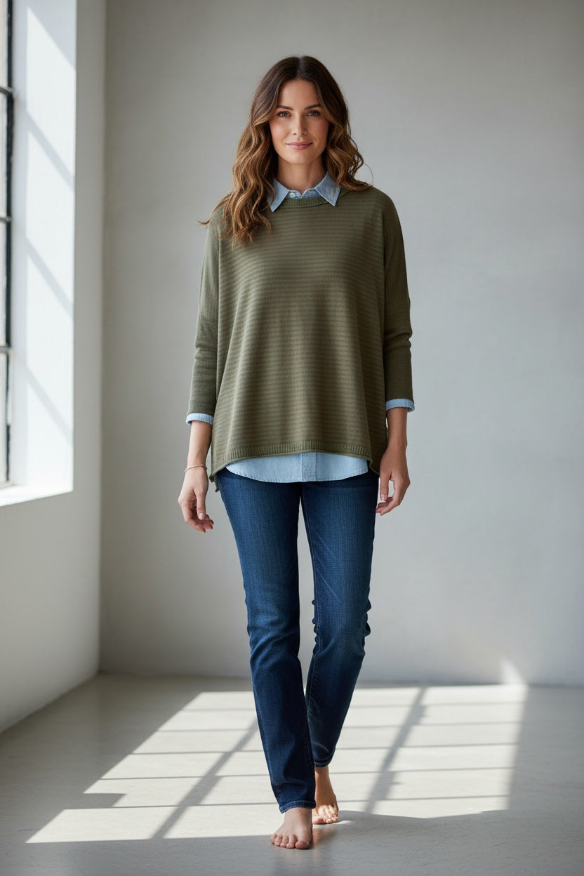 A woman with wavy brown hair stands barefoot on a sunlit floor near a window, smiling softly at the camera while wearing the MERSEA Catalina Crewneck Sweater in Jungle Stripe—an oversized green sweater by MERSEA layered over a blue shirt and paired with blue jeans.