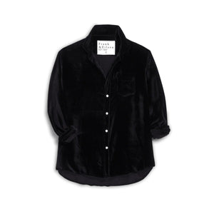 A smiling woman wears the FRANK & EILEEN EILEEN Relaxed Button Up in Black Italian Washed Velvet as she stands by a Christmas tree in a cozy, warmly lit living room, holding a glass of wine with bookshelves nearby.