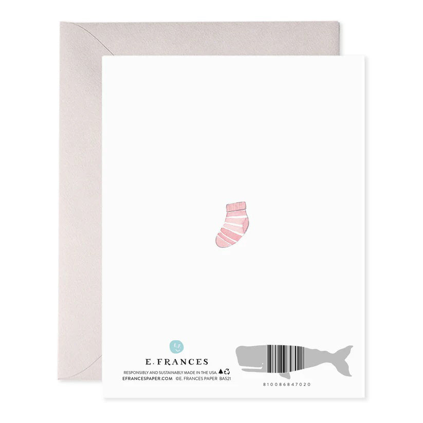 The E. FRANCES PAPER - BABY HEART GREETING CARD, by E. FRANCES PAPER, is handcrafted in the USA, featuring baby items like a rainbow, bib, bottle, bunny toy, onesie, stacking toys, pacifiers, and a wooden brush arranged in a heart shape with "hi baby!" at the center.