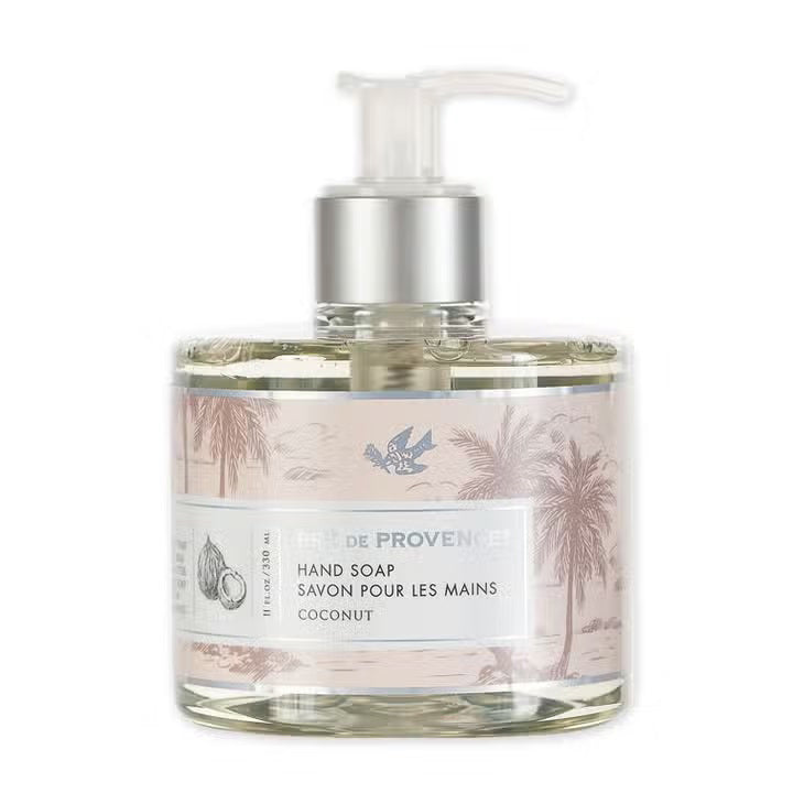 A clear pump bottle of liquid soap with an off-white label featuring an illustration of palm trees. The label reads "HERITAGE LIQUID SOAP - COCONUT" from "EUROPEAN SOAPS, LLC." A small drawing of a coconut is also visible on the label, evoking the charm of the French Riviera.