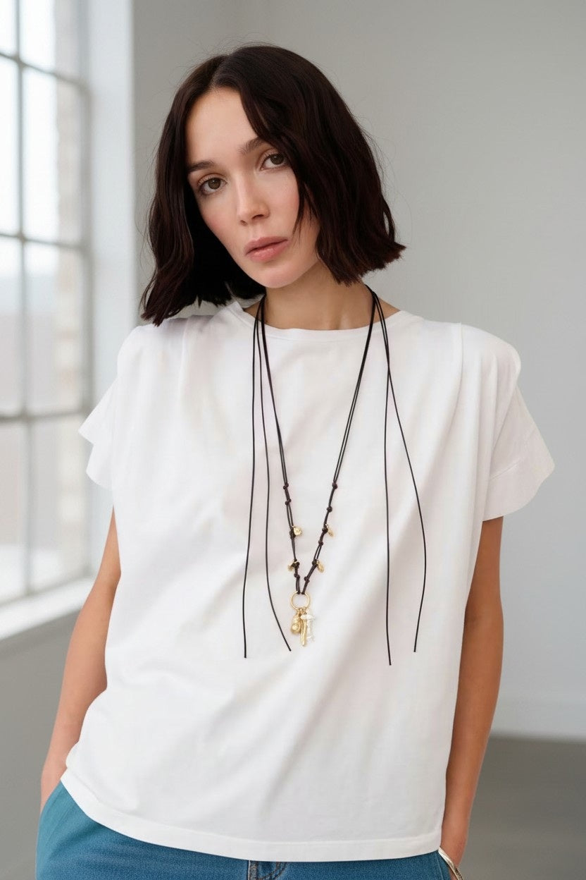A woman with short dark hair is seen standing in a bright room with large windows, wearing the FLOOR – WHITE COTTON TOP by FLOOR, styled in a modern silhouette with layered necklaces and blue jeans, looking directly at the camera.
