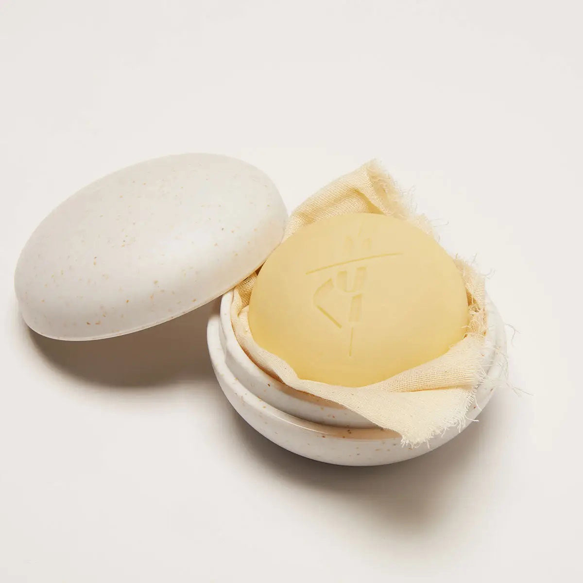 A KATE MCLEOD round yellow soap bar with an engraved symbol sits in the open KATE MCLEOD - THE FULL SIZE TRAVEL CASE, a speckled cream ceramic container with a lid on the side, ideal for eco-friendly travel.