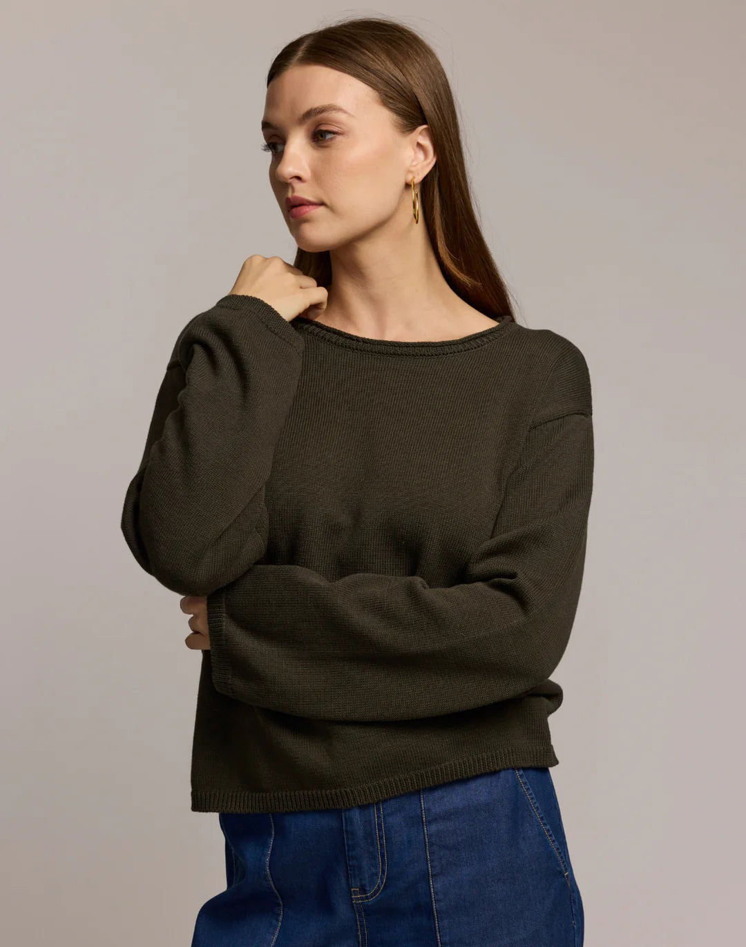 A woman with long brown hair and large hoop earrings models the HINSON WU AUDREY Long Sleeve Cotton Silk Sweater in tan with blue jeans, standing against a plain beige background, one hand raised near her shoulder.