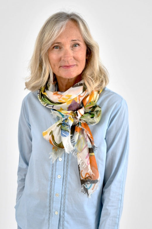 A smiling older woman with shoulder-length blonde hair wears a light blue button-up shirt and the STORIATIPIC - LOVE MAX SCARF IN WHITE by STORIATIPIC, standing against a plain white background.