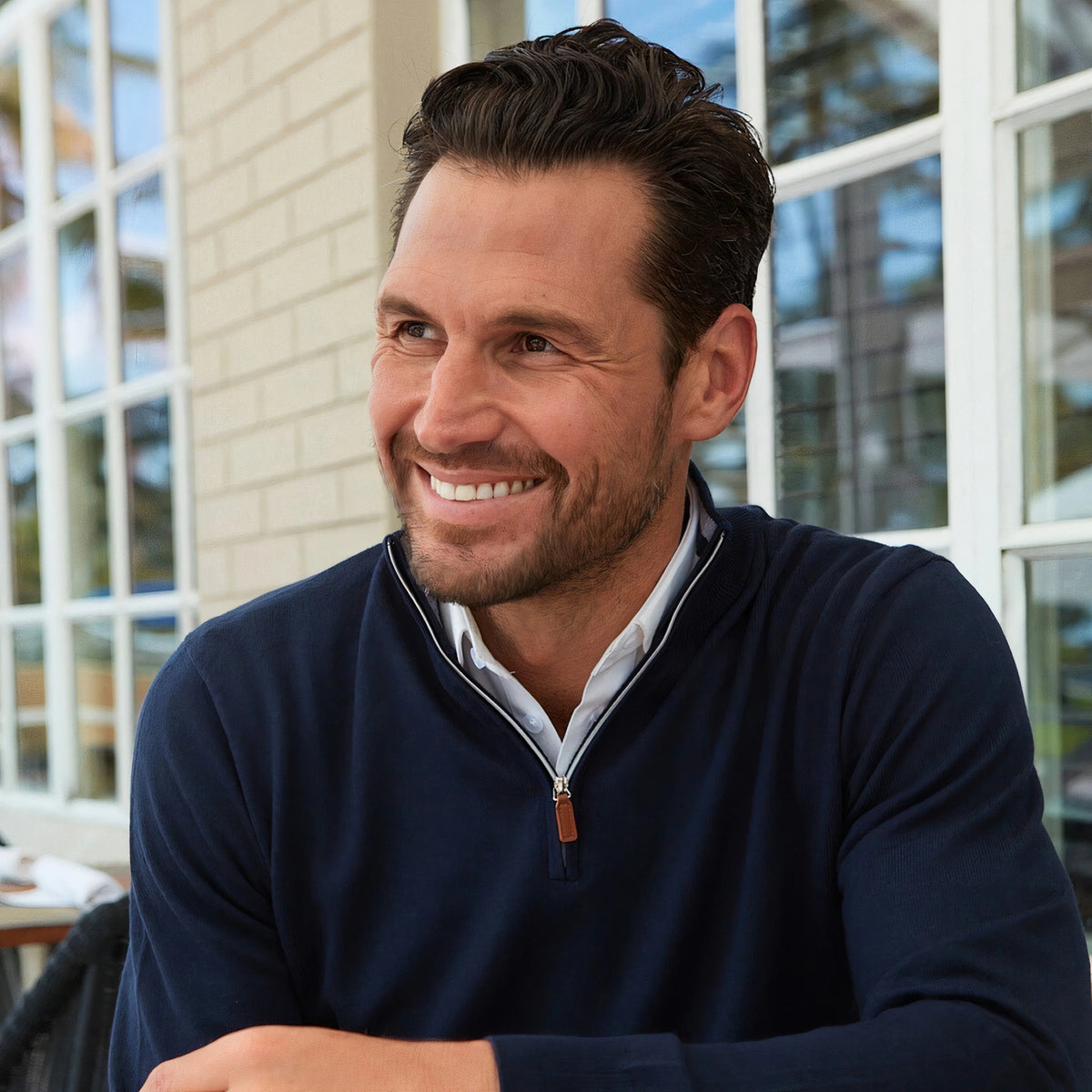 A man wearing the COLLARS AND CO Cashmere Touch Australian Merino Wool 1/4 Zip Sweater in Navy over a white shirt stands in front of open wooden doors, gazing thoughtfully to the side. Warm indoor lighting in the background accentuates the refined quality of this classic men’s layering piece from COLLARS AND CO.