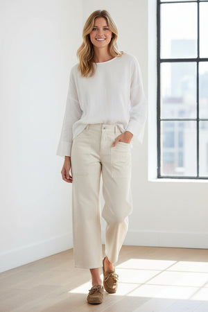 A person wearing FRANK & EILEEN’s Bray Everyday Barrel Pant in Italian Peached Twill Chalk pairs them with a white long-sleeve top, standing casually with one hand in their pocket.