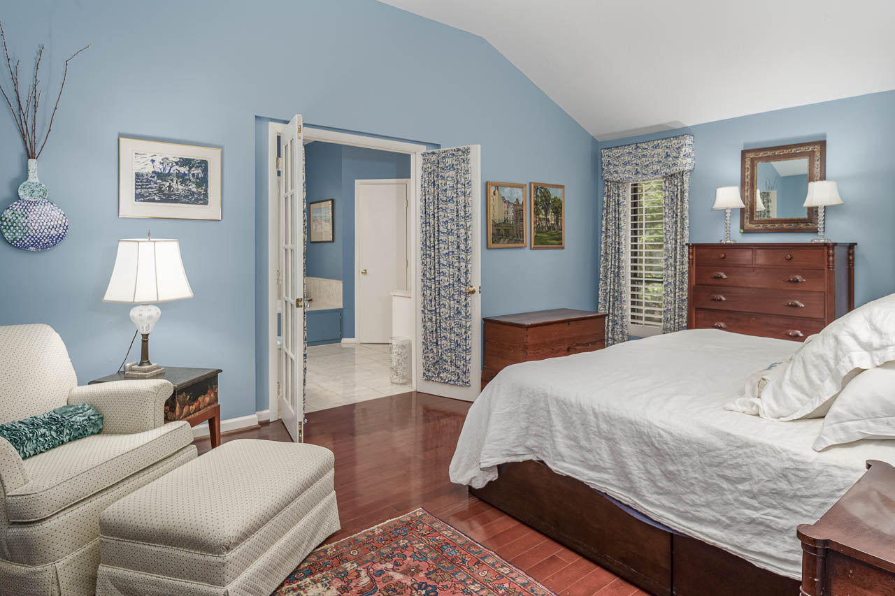 A cozy bedroom with pale blue walls, white bedding, wooden furniture, an armchair with ottoman, a colorful rug, and decorative artwork. The room opens to a bathroom with blue and white patterned curtains.