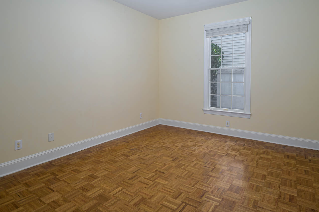 An empty room with light yellow walls, a parquet wood floor, white baseboards, and a single window with white blinds partially open, letting in natural light.