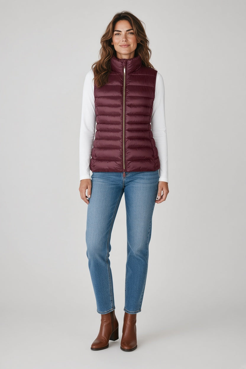 A woman wearing a dark red SAVE THE DUCK - LYNN PUFFER VEST IN WINE with a standing collar over a white long-sleeved shirt, blue jeans, and brown ankle boots.