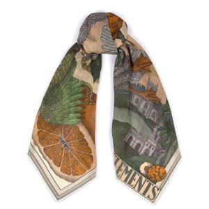 A person elegantly stands against a plain background wearing the SABINA SAVAGE ORANGES AND LEMONS 135CM cashmere scarf in Citrine/Zest, featuring animals, leaves, and objects that contrast beautifully with their light-colored dress.