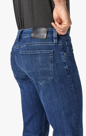 A man stands facing forward, wearing a dark short-sleeve polo, 34 HERITAGE - COURAGE STRAIGHT LEG JEAN IN MID URBAN WASH, a black belt, and black shoes. One hand is in his pocket; he has short hair, a trimmed beard, and a neutral expression.