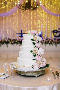 A three-tiered white wedding cake decorated with pink and white flowers sits on an ornate stand. Champagne glasses, utensils, and candles are arranged on the table, with golden lights and floral decor in the background.