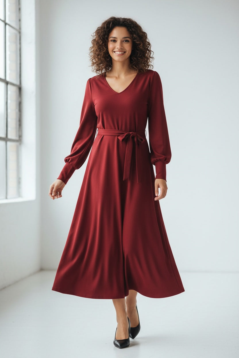 A woman with long, light brown hair stands against a plain background. She is wearing the SFIZIO long sleeve v-neck flare skirt dress, featuring a tie waist. Looking to her left with a calm expression, she has hoop earrings and exudes minimalist style.