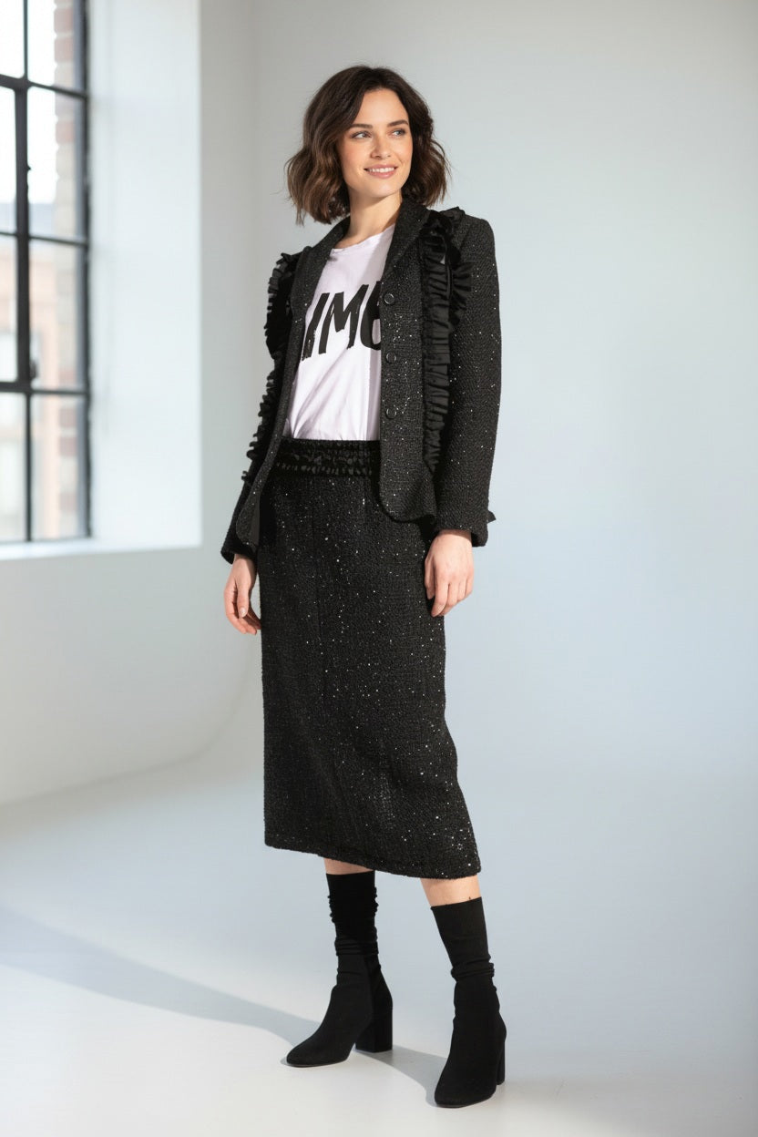 A woman wears the MEIMEIJ LUREX TWEED SKIRT with a black textured ruffle blazer, a white tee with black text, and black shoes with matching socks, posed against a white background.
