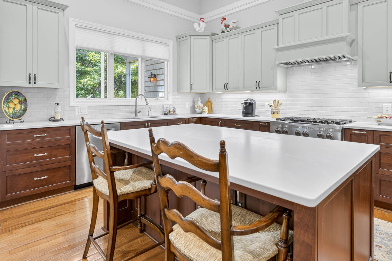 Bright kitchen with white cabinets, dark wood lower cabinets, a large island with two wooden chairs, white countertops, a gas stove, and a window with a view of greenery.