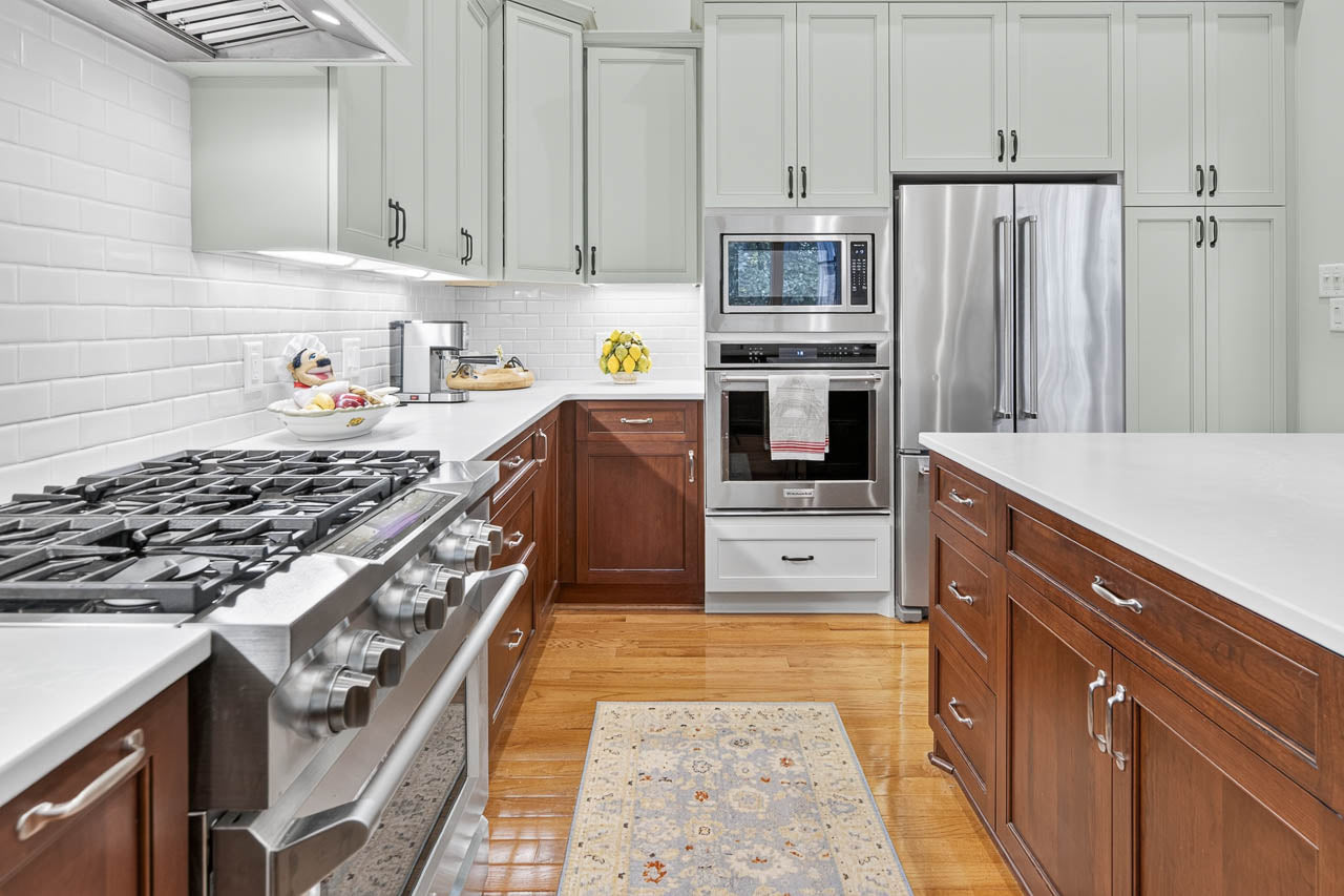 Modern kitchen with stainless steel appliances, white countertops, light and dark wood cabinets, fruit bowls, a gas stove, built-in microwave and oven, and a patterned rug on a wooden floor.