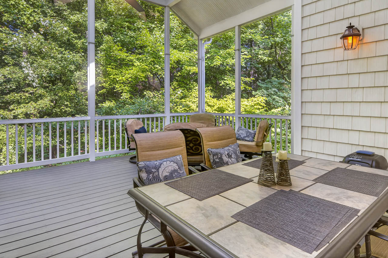 Screened-in porch with a tile-top dining table, cushioned chairs, and decorative placemats, surrounded by trees and greenery, with soft sunlight illuminating the space.