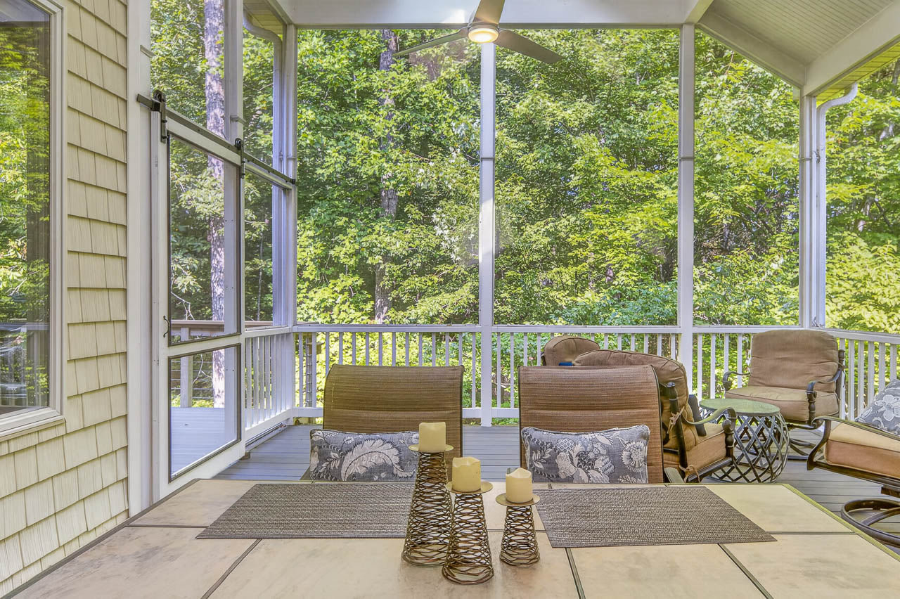 Screened-in porch with cushioned chairs, a table set with candles, and views of lush green trees outside. Ceiling fan overhead and outdoor seating area visible in the background.