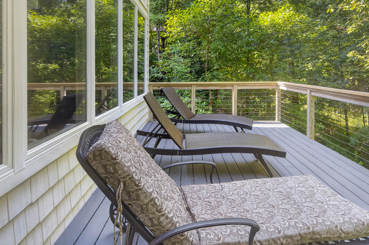 Three lounge chairs with patterned cushions are arranged on a wooden deck, next to a house with large windows, overlooking a lush, green forest.