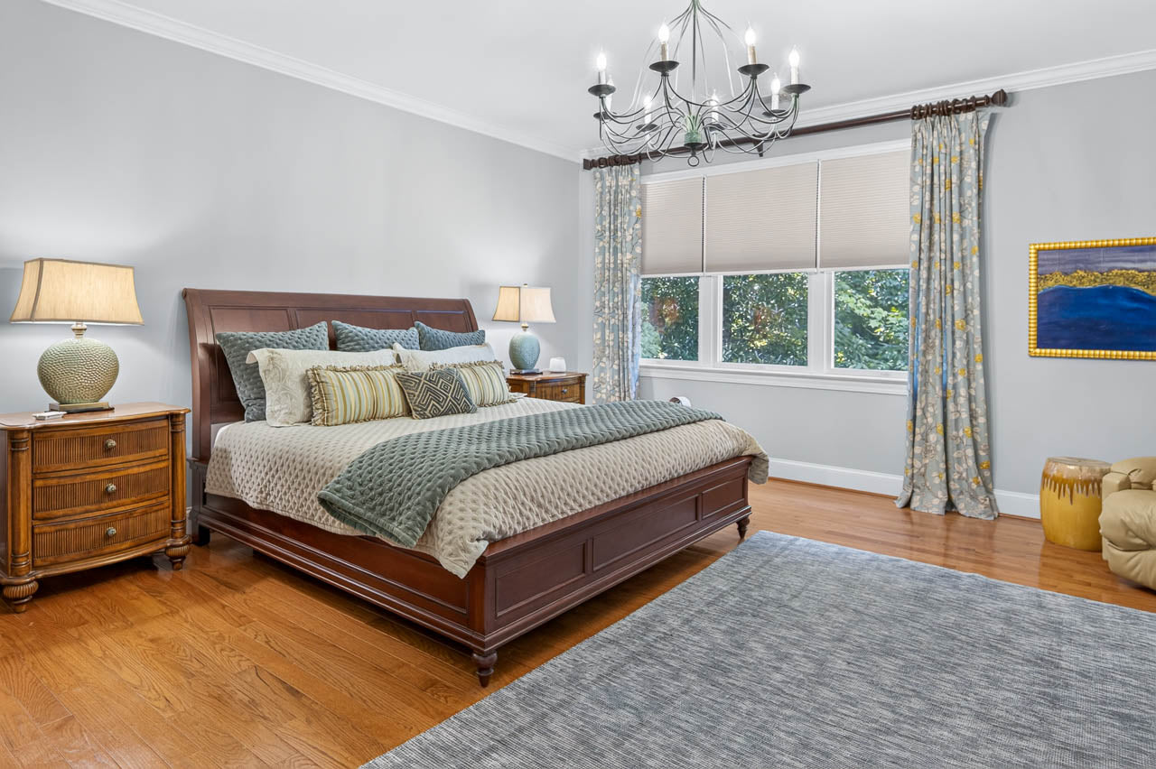Spacious bedroom with a wooden bed, gray bedding, two nightstands with lamps, a chandelier, large window with curtains, wood floor, and a blue-gray rug.