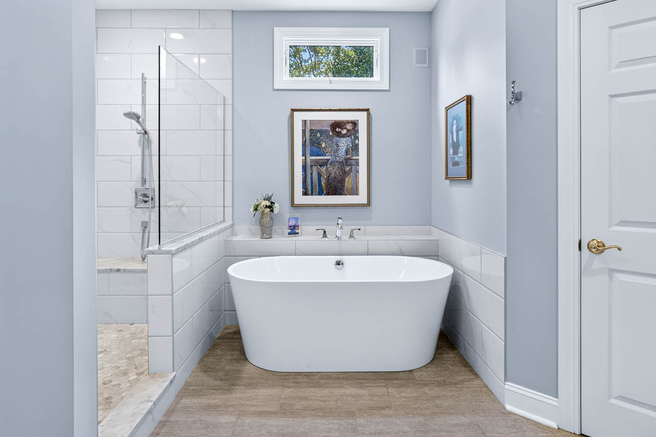Modern bathroom with a white freestanding tub, glass-enclosed shower, light gray walls, framed artwork above the tub, and a small window letting in natural light.