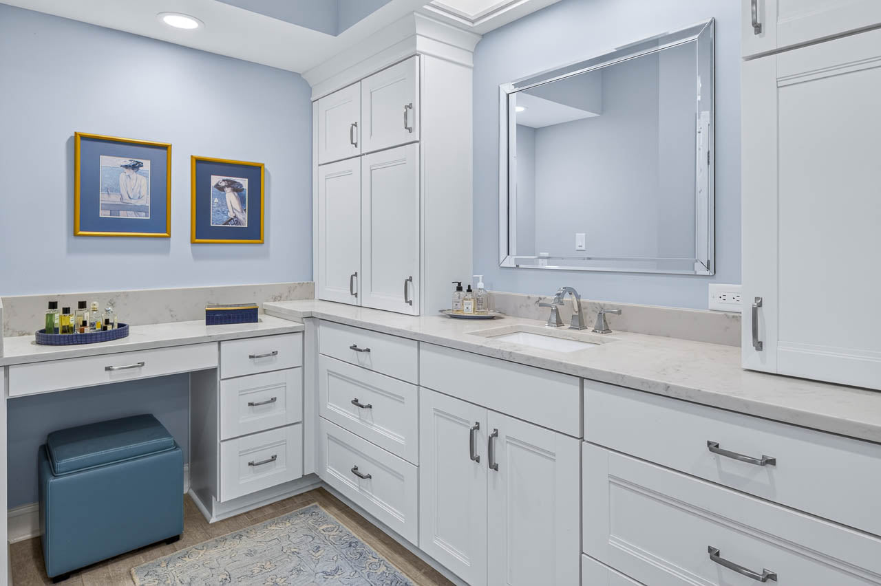 Modern bathroom with light blue walls, white cabinets, marble countertop, square mirror, two framed bird prints, blue ottoman, soap dispenser, and toiletries neatly arranged on the counter.