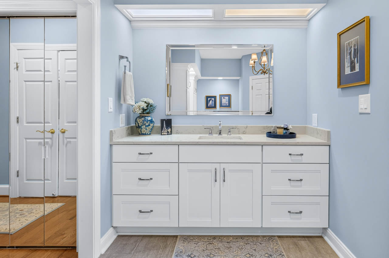 A bathroom with a white vanity, large mirror, blue walls, decorative flowers, towel, and framed photos. There is a mirrored closet door on the left and a rug on the wooden floor.