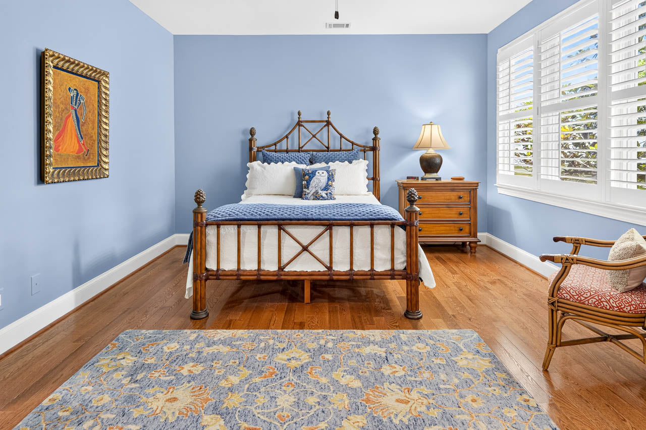 A cozy bedroom with a wooden bed, blue walls, floral rug, wooden dresser with a lamp, chair, and large window with white shutters letting in natural light. A colorful painting hangs on the left wall.