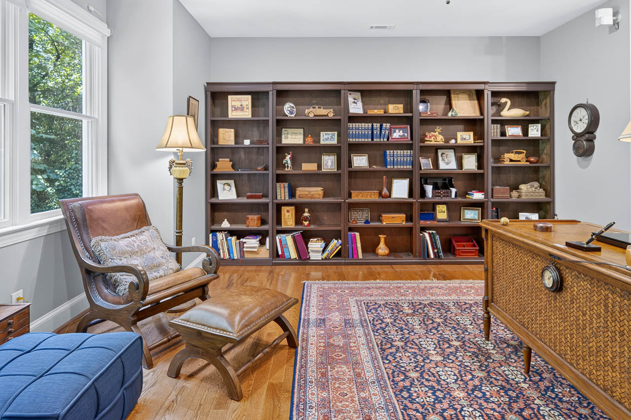 A cozy home office with a wooden desk, leather chair, ottoman, floor lamp, and large bookshelf filled with books, framed photos, and decorative items. A window lets in natural light. Rug and hardwood floors complete the room.