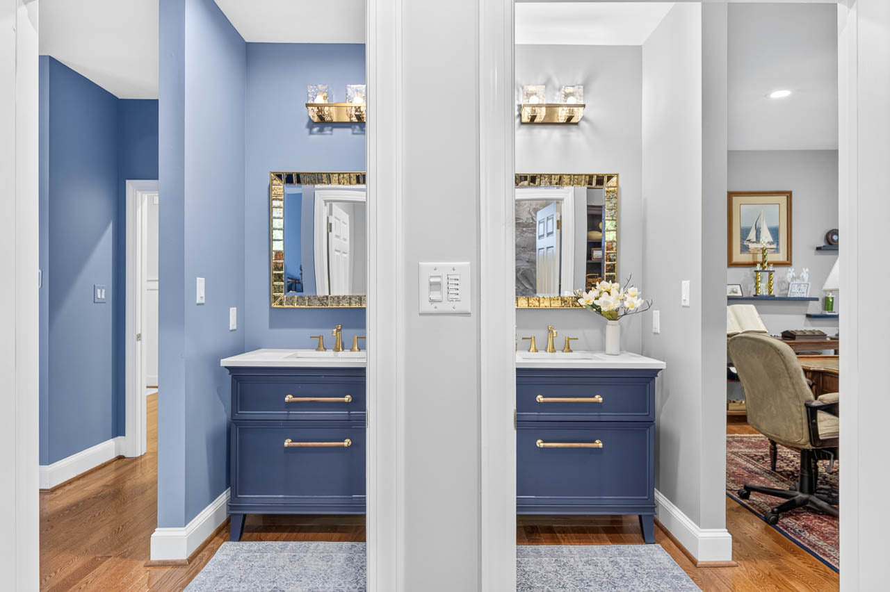 Two blue bathroom vanities with gold fixtures and matching mirrors are separated by a wall. To the right, an office area with a desk and chair is partially visible. The floors are wood, and the walls are painted blue and white.