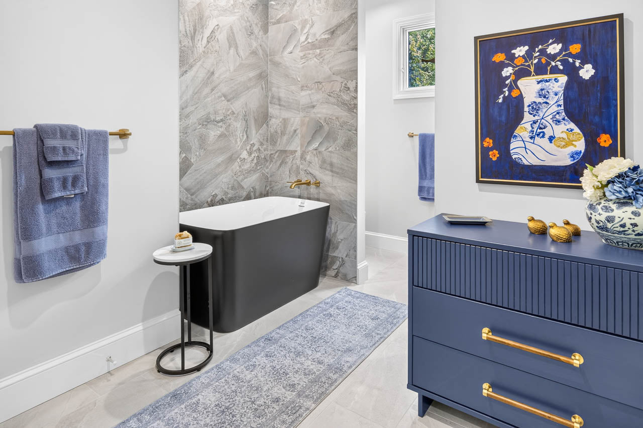 Modern bathroom with a black tub, marble tile wall, blue towels, blue dresser with gold handles, floral art, small round table, and window. Decor includes a vase with flowers and gold bird figurines.
