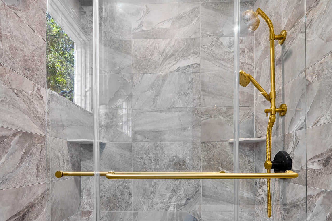 Modern shower with gray marble tiles, a gold showerhead, matching gold grab bar, and a small window letting in natural light. The design is sleek and accessible.