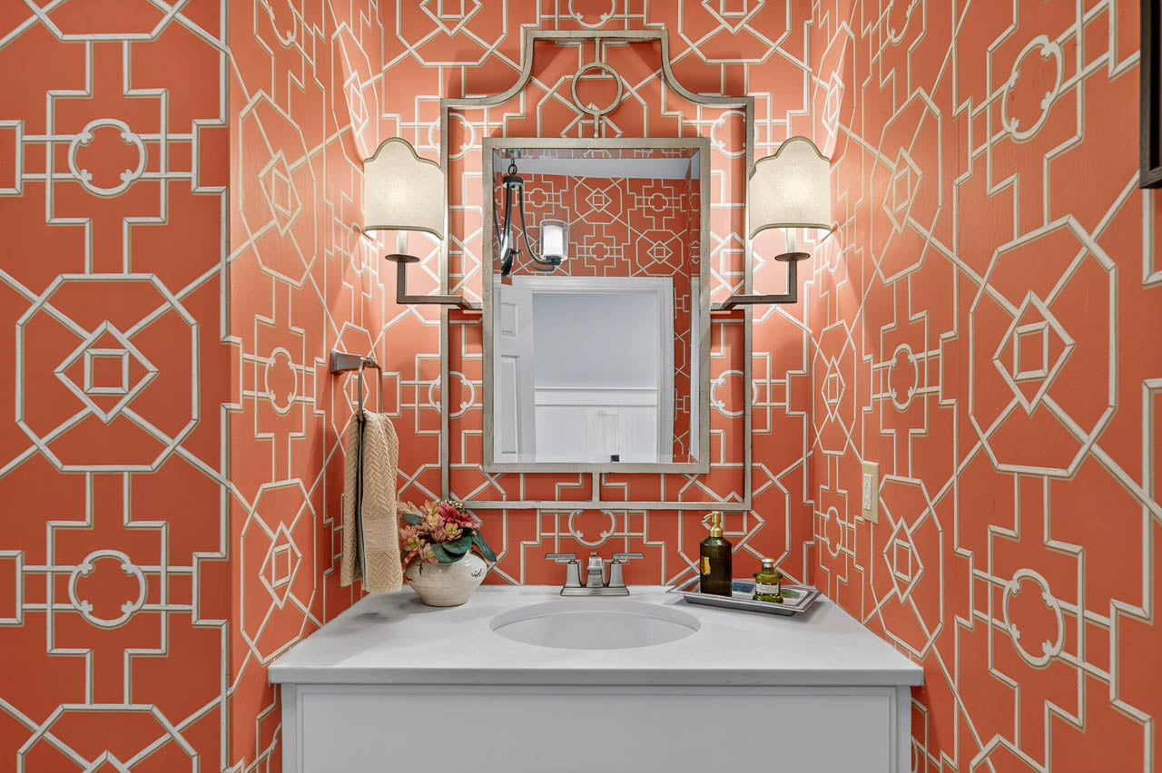 Small bathroom with bold orange geometric wallpaper, white vanity and sink, two wall sconces, a towel hanging on the left, a mirror above the sink, and decorative items arranged on the countertop.