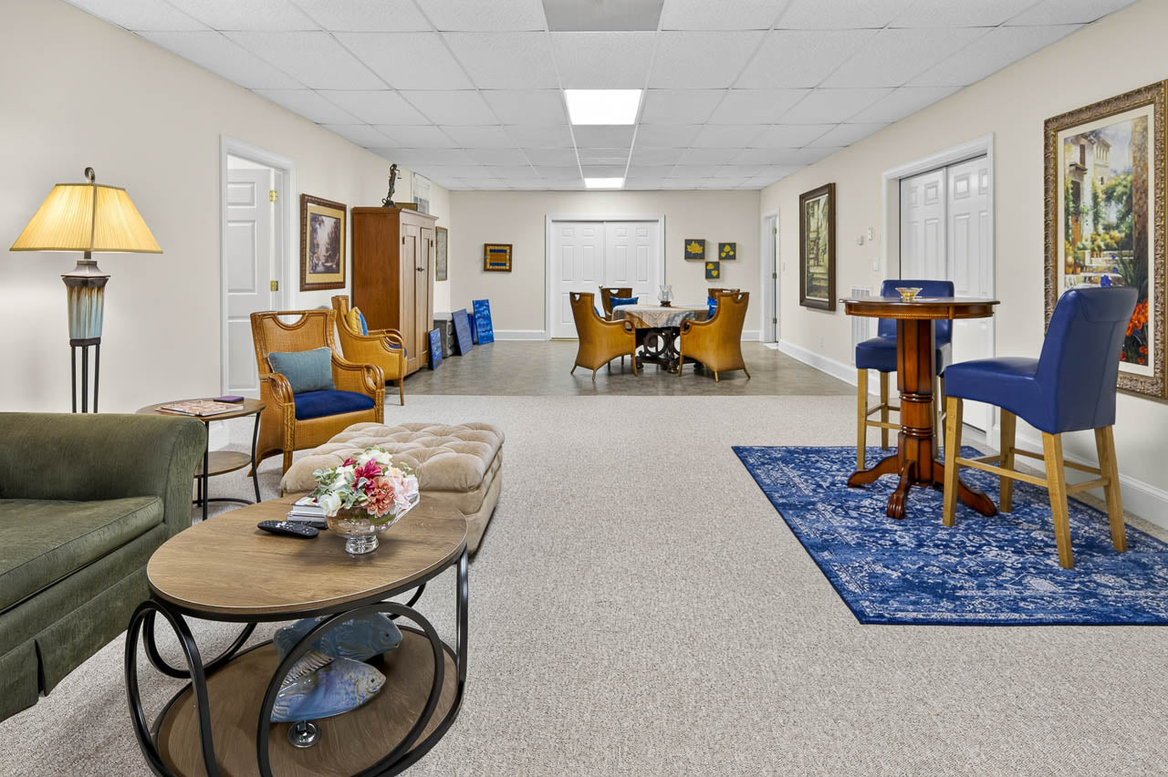 A spacious, well-lit lounge with various chairs, tables, floral decor, framed art on cream walls, and blue accents on rugs and chairs. The room has a cozy, welcoming atmosphere.